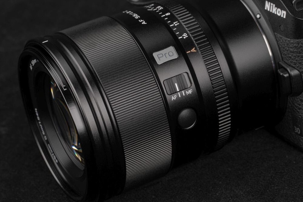 Czarny obiektyw z widocznymi oznaczeniami \'Pro\', \'AF 56mm f/1.2\', oraz przełącznikiem \'AF/MF\'. W tle częściowo widoczny aparat z logo \'Nikon\'.