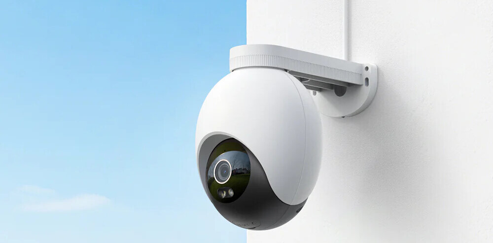 Na obrazie widoczna jest zewnętrzna kamera obrotowa zamontowana na białej ścianie. Kamera ma biało-czarną obudowę i jest skierowana w stronę otwartej przestrzeni z niebieskim niebem.