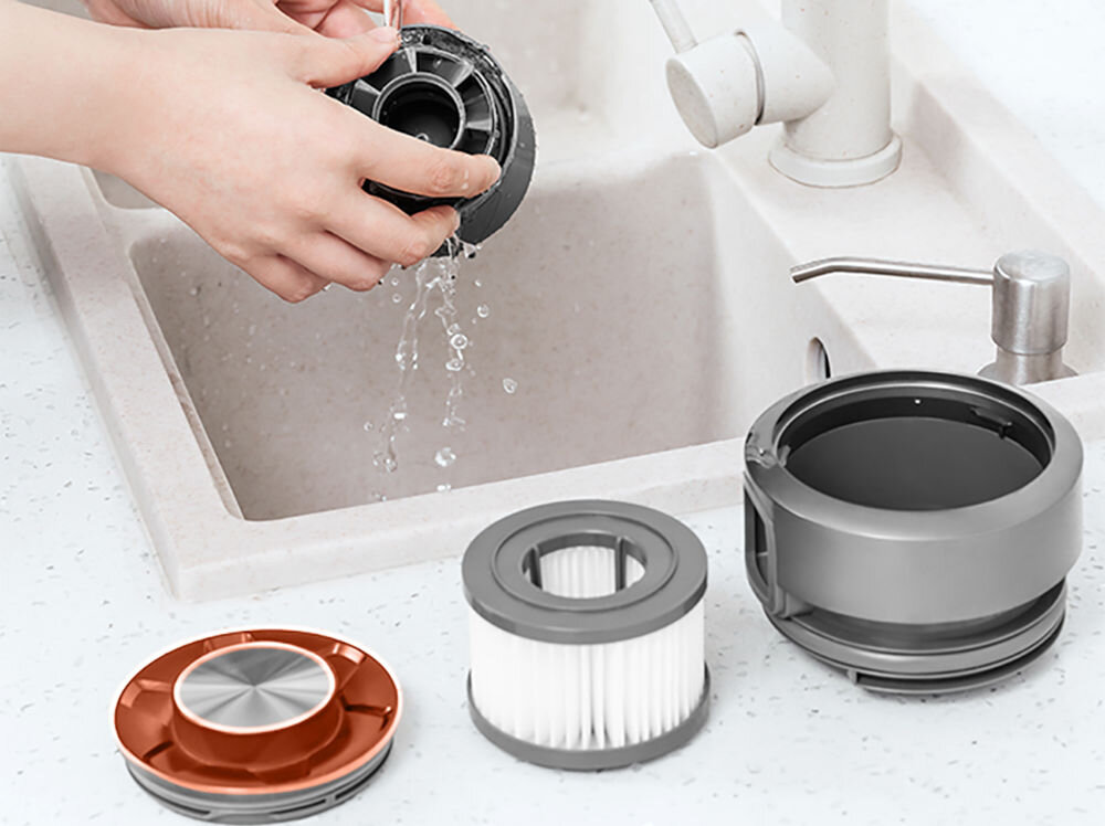 Osoba myje część odkurzacza pod bieżącą wodą w zlewie. Obok znajdują się trzy inne części odkurzacza, w tym filtr i elementy obudowy.