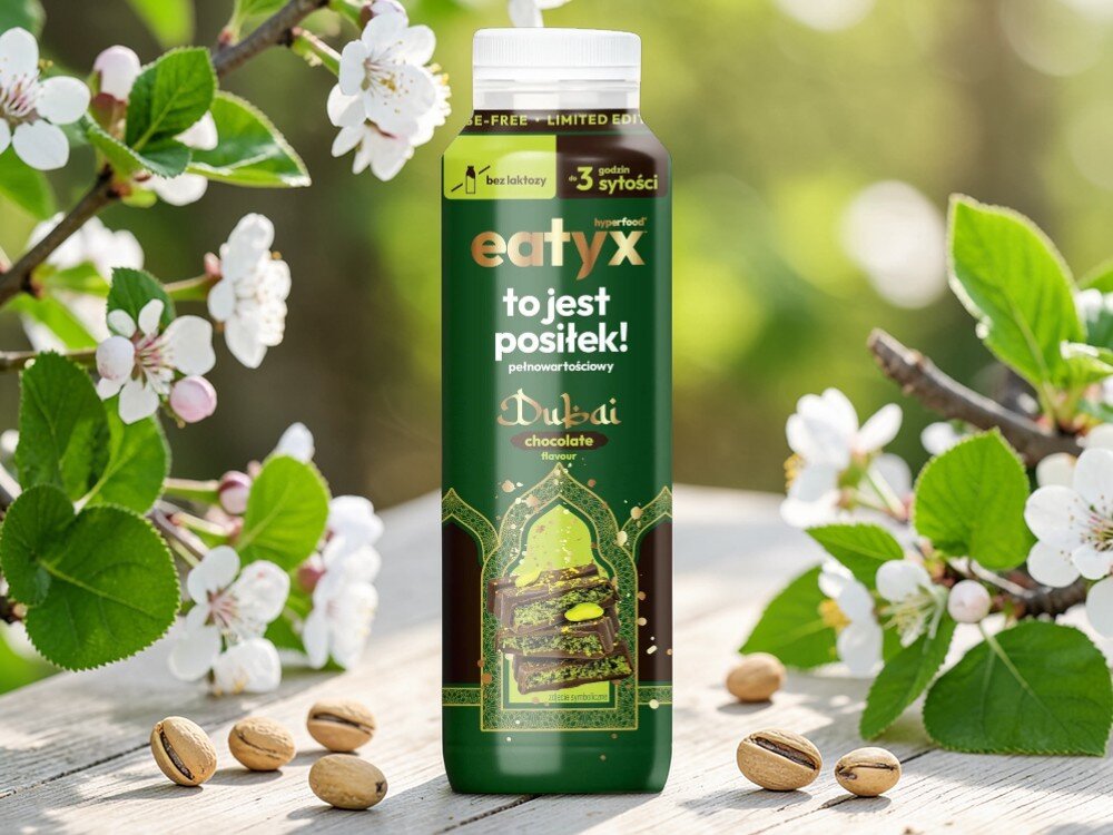 Posiłek w płynie EATYX Hyperfood Dubajska Czekolada (300 ml) butelka posiłku stoi na drewnianej podłodze obok leżą pistacje białe kwiatki 23 witamin i minerałów biotyna (10,2 µg) witamina B12 (0,84 µg) chrom (6,6 µg) selen (9,3 µg) witamina E (5,4 g) wapń (540 mg) wspierają zdrowie skóry, włosów, paznokci i metabolizmu energetycznego