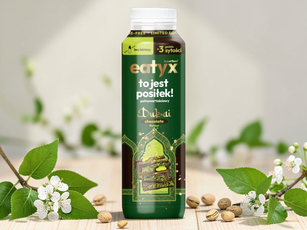 Posiłek w płynie EATYX Hyperfood Dubajska Czekolada (300 ml) butelka posiłku stoi na drewnianej podłodze obok leżą pistacje białe kwiatki olej słonecznikowy źródło kwasów omega-6 i witaminy E chroni komórki przed stresem oksydacyjnym oraz wspiera serce, skórę i odporność węglan wodorotlenku magnezu magnez niezbędny dla kości, mięśni i układu nerwowego