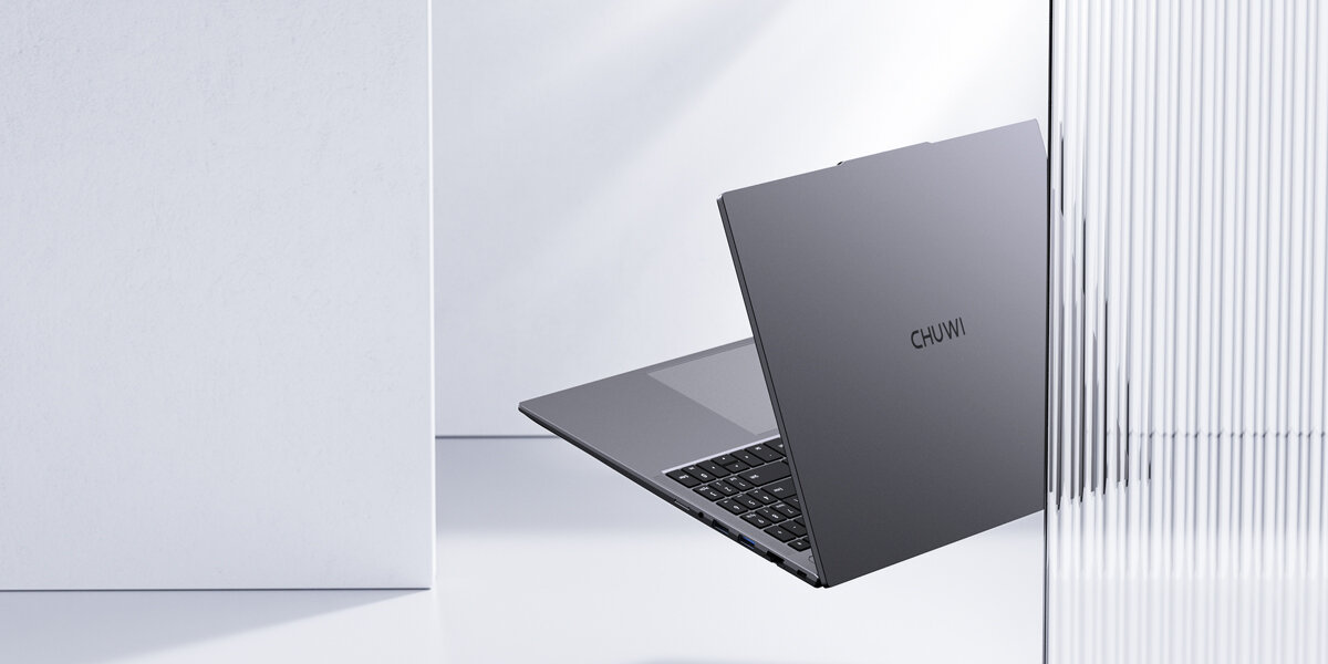 Laptop w kolorze szarym z widocznym logo CHUWI na pokrywie, ustawiony w minimalistycznym, jasnym otoczeniu z elementami szklanymi i betonowymi.