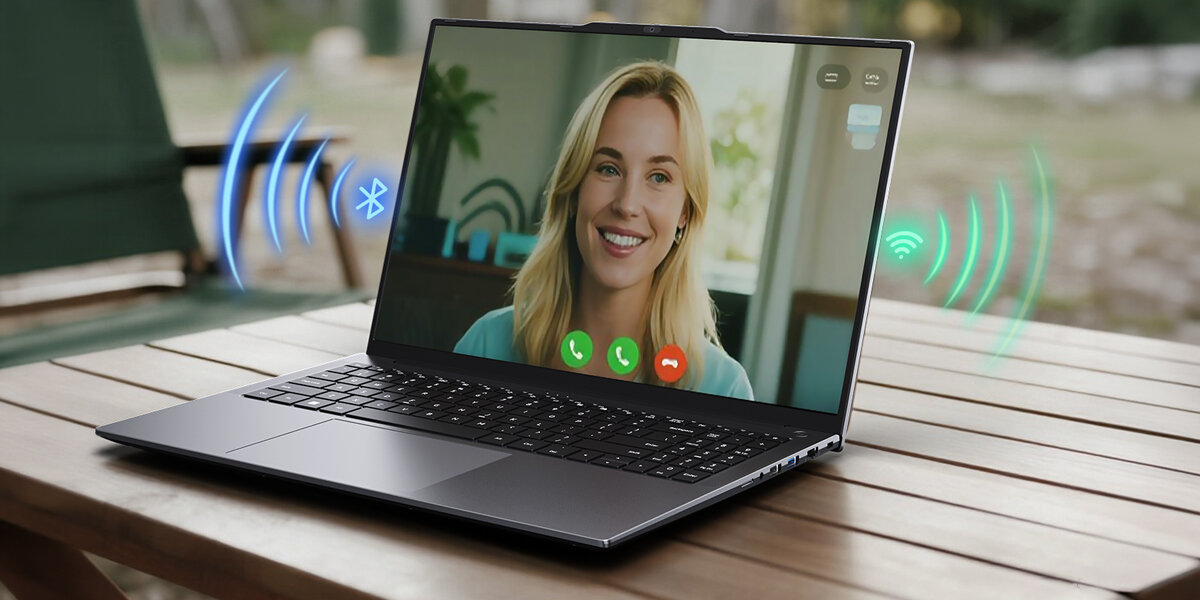 Na drewnianym stole stoi otwarty laptop z wyświetlonym obrazem kobiety podczas wideorozmowy. Obok laptopa widoczne są ikony Bluetooth i Wi-Fi.