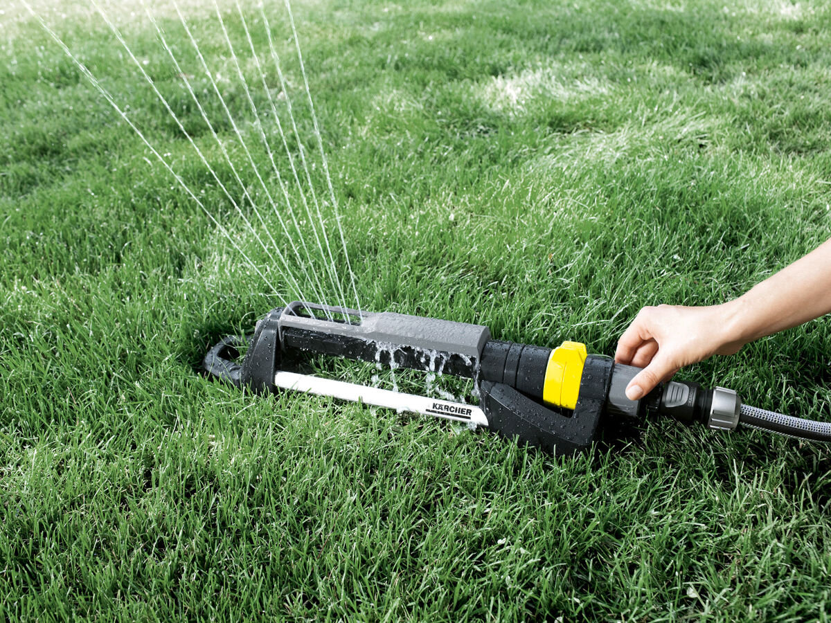 Zraszacz KARCHER OS 5.320 S, przesłona, SplashGuard, chlapanie, regulacja, w tle trawnik, na nim zraszacz i dłoń obsługująca przesłonę SplashGuard
