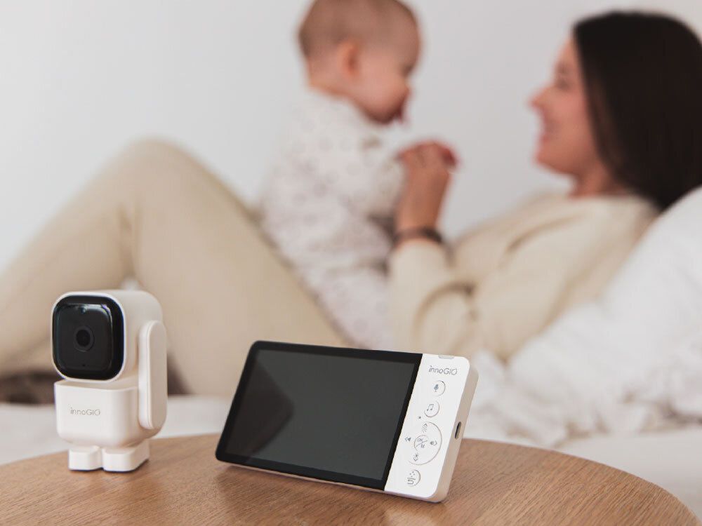 Na drewnianym stole znajduje się elektroniczna niania z kamerą i monitorem. W tle widoczna jest osoba dorosła trzymająca dziecko.