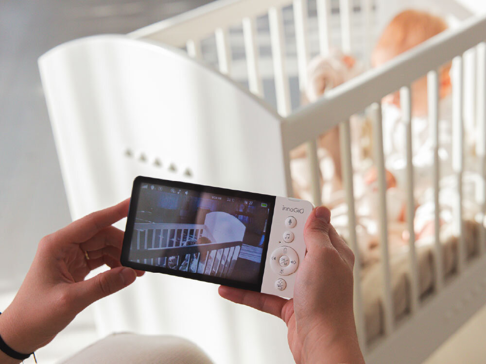 Osoba trzyma urządzenie z ekranem, na którym widać obraz łóżeczka dziecięcego. W tle znajduje się niemowlę w białym łóżeczku, a na urządzeniu widoczne jest logo \'innaGIO\'.