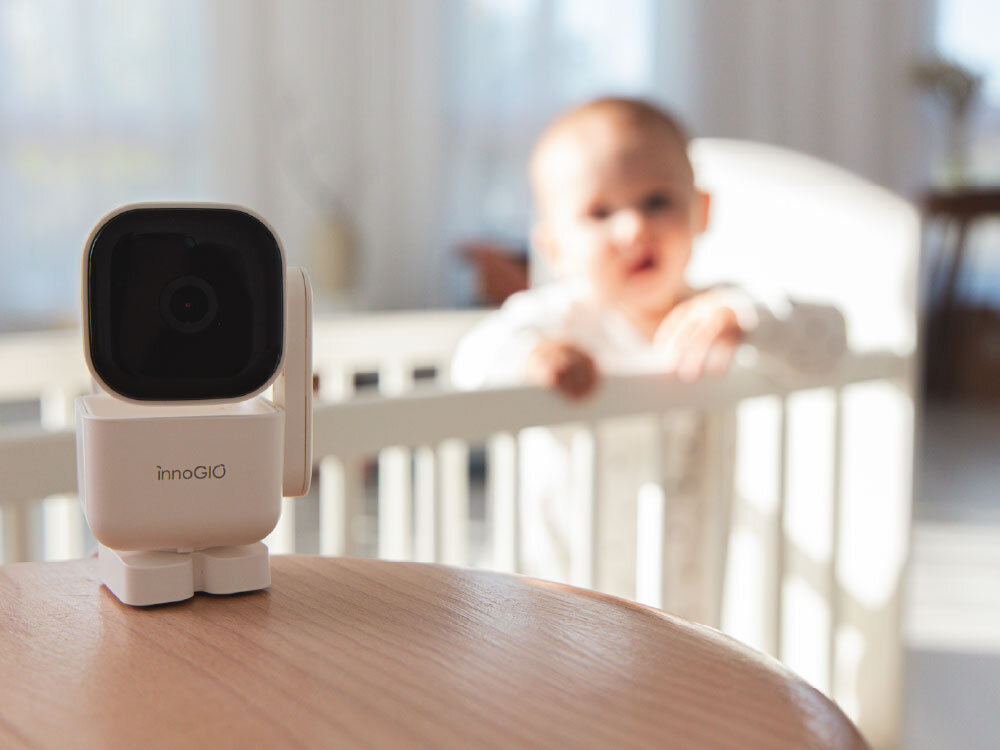 Na drewnianym stole stoi elektroniczna niania z logo \'innoGIO\'. W tle widać dziecko w łóżeczku.