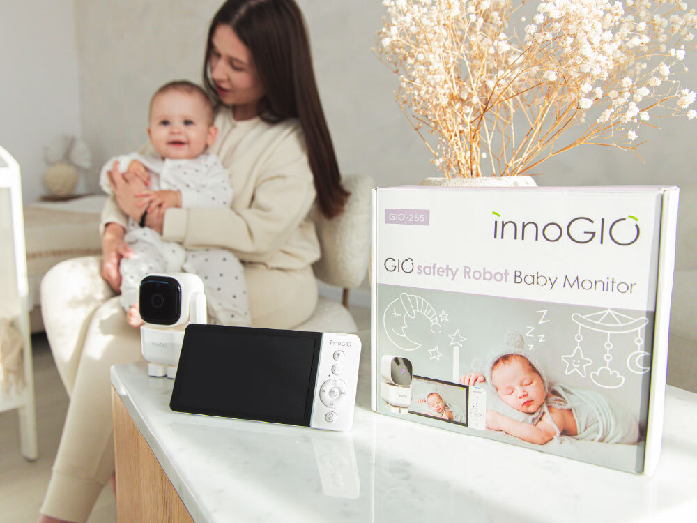 Na stole znajduje się elektroniczna niania z ekranem i kamerą, obok pudełko z napisem \'innoGIO GIO safety Robot Baby Monitor\'. W tle kobieta trzymająca dziecko na kolanach.
