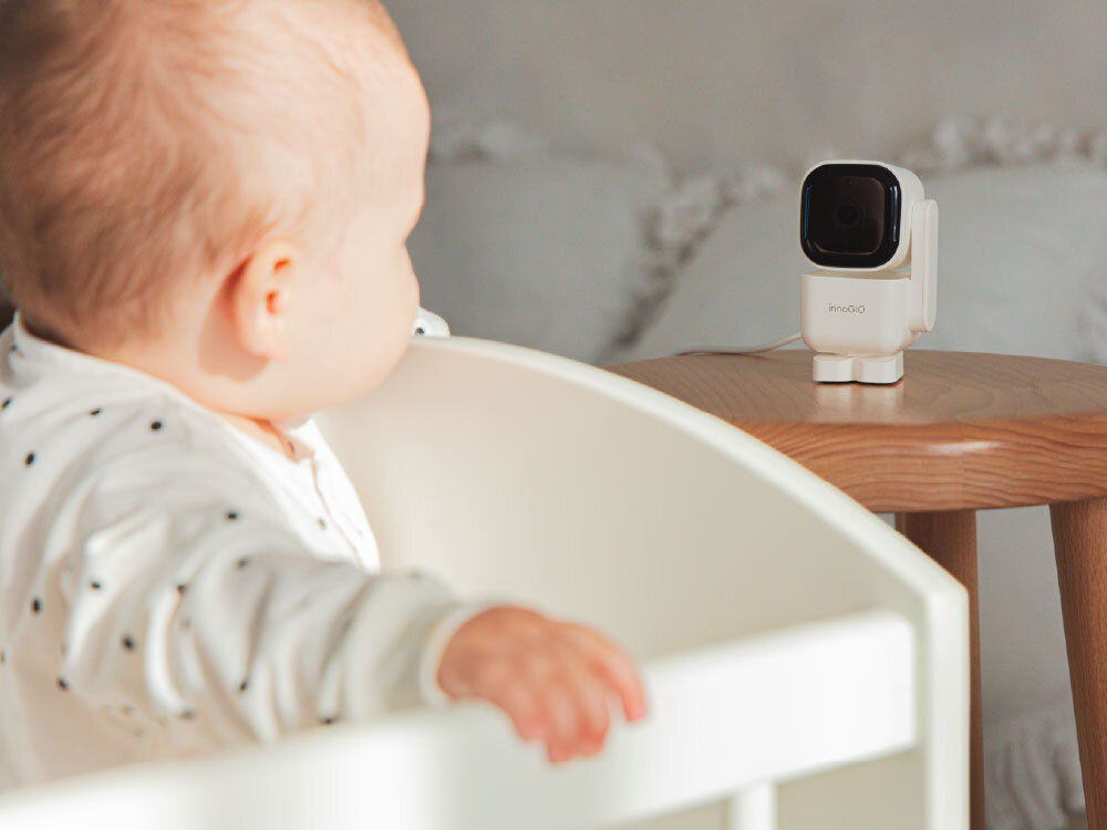 Na drewnianym stoliku stoi elektroniczna niania z napisem \'innogio\'. W tle widoczny jest niemowlak w białym łóżeczku.