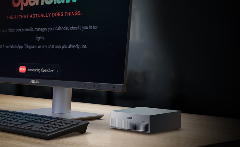 Komputer ASUS Ascent GX10 Ustawiony na drewnianym biurku srebrny komputer typu mini PC, obok czarnej klawiatury i u dołu czarnego monitora wyświetlającego ciemną tapetę ze świecącym czerwonym przyciskiem 