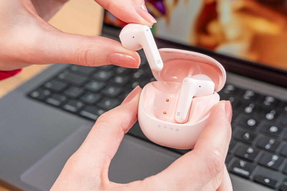 Słuchawki douszne XMUSIC XTW300P różowy Dłonie wyjmują różową słuchawkę douszną z otwartego, czarnego etui. Słuchawki znajdują się nad klawiaturą laptopa.