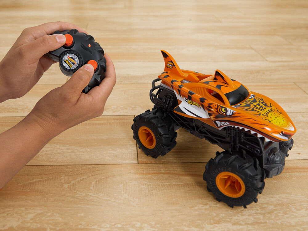 Samochód zdalnie sterowany Hot Wheels Tiger Shark HNV03 Dwie dłonie trzymające czarny kontroler (pilot) do zdalnego sterowania, z pomarańczowymi przyciskami. Obok, na drewnianej podłodze, stoi zdalnie sterowany samochód Hot Wheels Monster Truck Shark w pomarańczowo-czarnym malowaniu z motywem tygrysa/rekina.