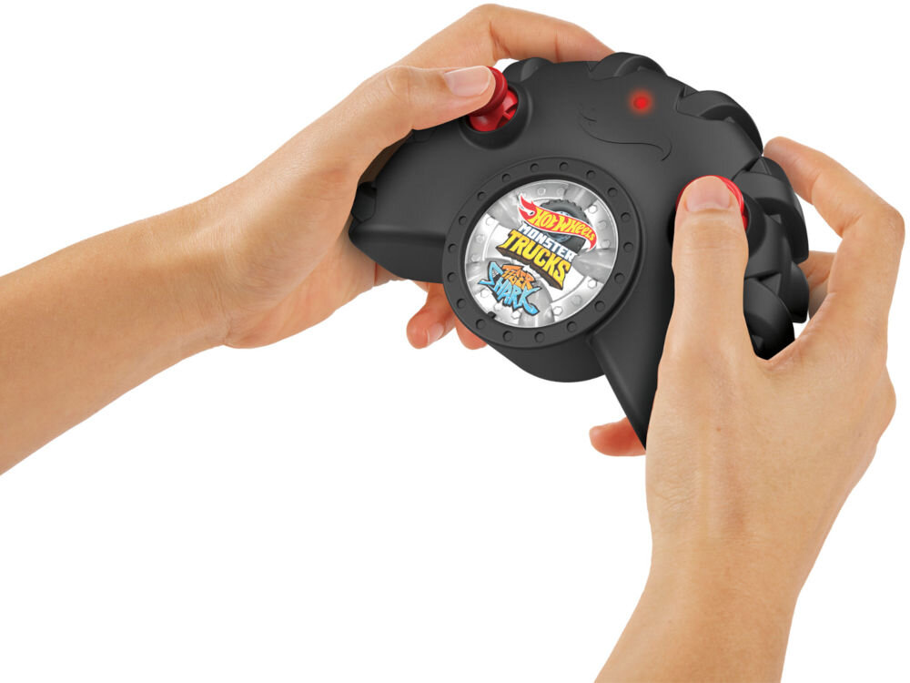 Samochód zdalnie sterowany Hot Wheels Tiger Shark HNV03 Zbliżenie na dwie dłonie trzymające czarny kontroler (pilot) do zdalnie sterowanego pojazdu. Na środku kontrolera widoczne jest logo Hot Wheels Monster Trucks Shark. Tło jest białe.