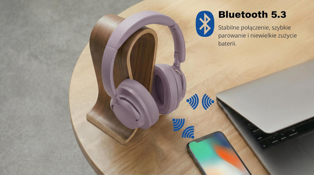 Różowe słuchawki nauszne umieszczone na drewnianym stojaku na stole obok smartfona i laptopa. Obok widoczny jest tekst: \'Bluetooth 5.3 Stabilne połączenie, szybkie parowanie i niewielkie zużycie baterii.\'