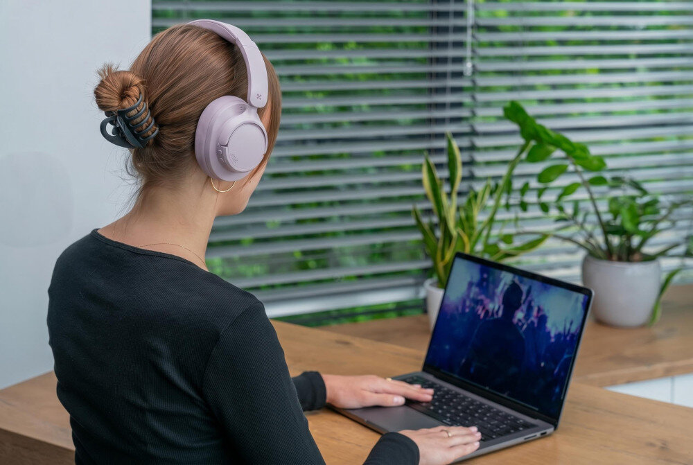 Osoba siedzi przy drewnianym stole, nosząc różowe słuchawki nauszne i pracując na laptopie. W tle widać rośliny doniczkowe i żaluzje na oknie.