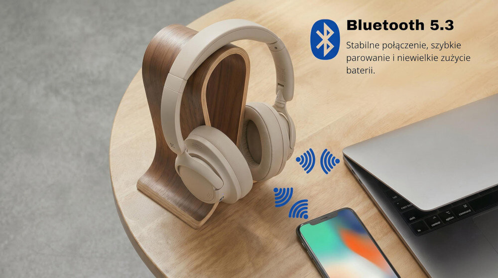 Na drewnianym stojaku znajdują się kremowe słuchawki nauszne obok laptopa i smartfona. Obok widnieje tekst: \'Bluetooth 5.3 Stabilne połączenie, szybkie parowanie i niewielkie zużycie baterii\'.