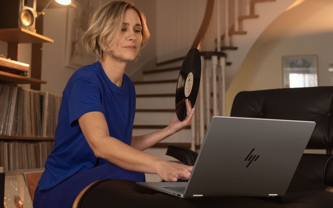 Kobieta w niebieskim stroju korzysta z laptopa HP OmniBook w salonie, w ręku trzyma płytę winylową, na półkach kolekcja winyli.