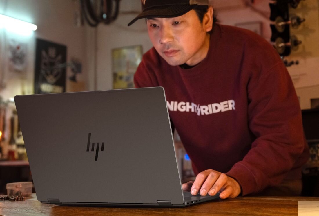 Praca przy laptopie HP OmniBook w klimatycznym warsztacie, mężczyzna w bordowej bluzie korzysta z komputera.