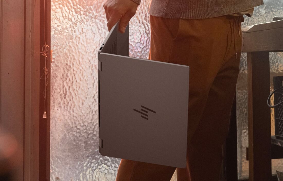 Przenoszenie otwartego laptopa HP OmniBook przy drzwiach z matowym szkłem w ciepłym świetle.