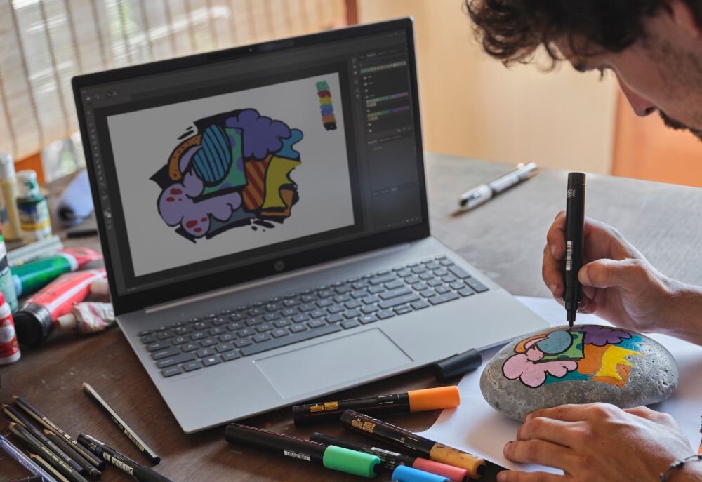 Laptop HP OmniBook podczas procesu projektowania graficznego, użytkownik maluje kolorowy wzór na kamieniu, widoczne akcesoria artystyczne na stole.