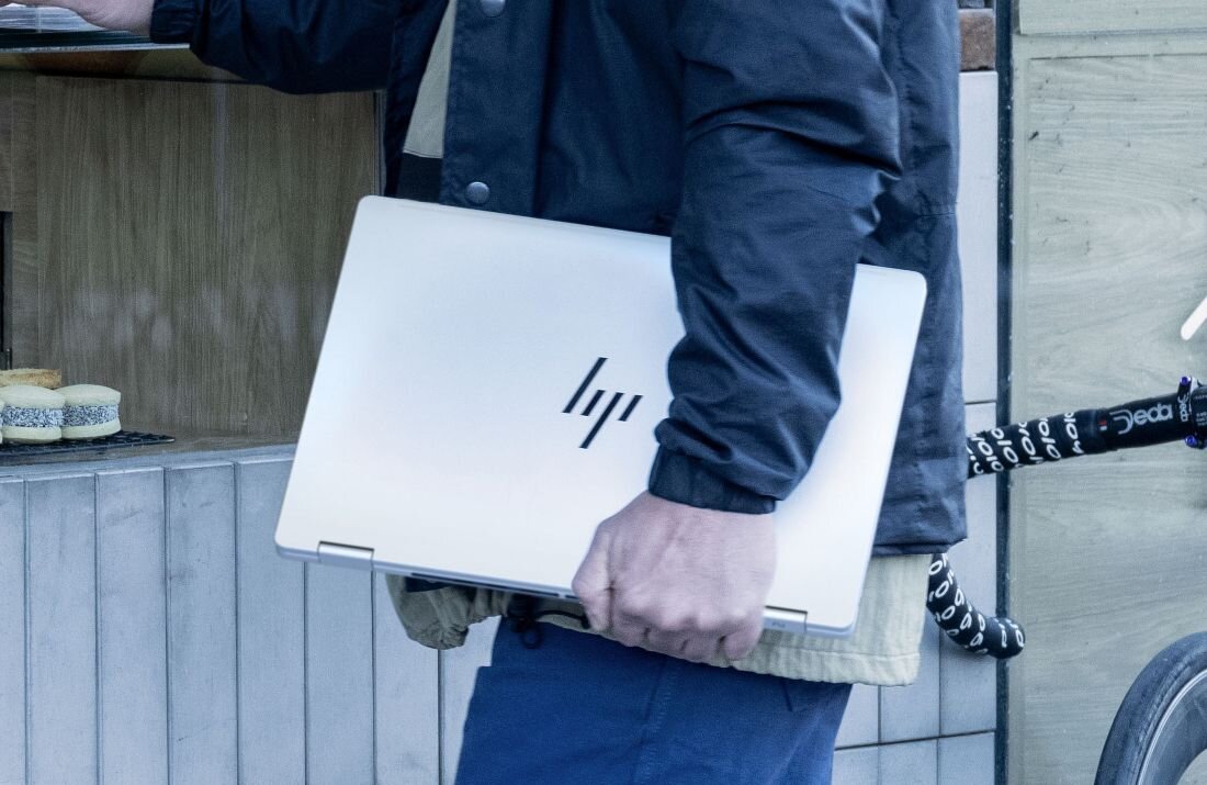 Mężczyzna trzymający zamknięty laptop HP OmniBook w pobliżu baru z ciastkami, rower oparty o ścianę.