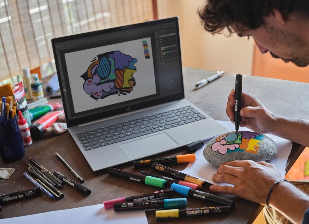 Projektant w pomarańczowej koszulce malujący kamienie markerami obok laptopa HP OmniBook 5 z grafikami na ekranie - Prezentacja laptopa HP OmniBook 5 16-BC1