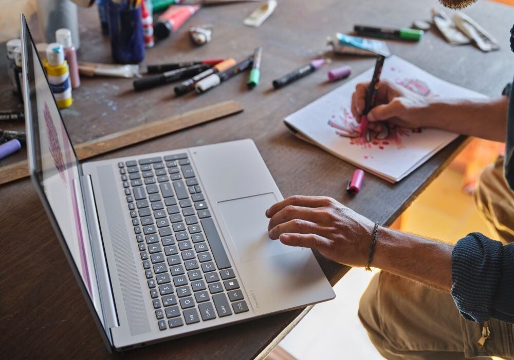 Ręka mężczyzny na klawiaturze laptopa HP OmniBook 5 16-BC1 podczas pracy kreatywnej z kolorowymi markerami na stole - Pełnowymiarowa klawiatura z białym podświetleniem LED i blokiem numerycznym