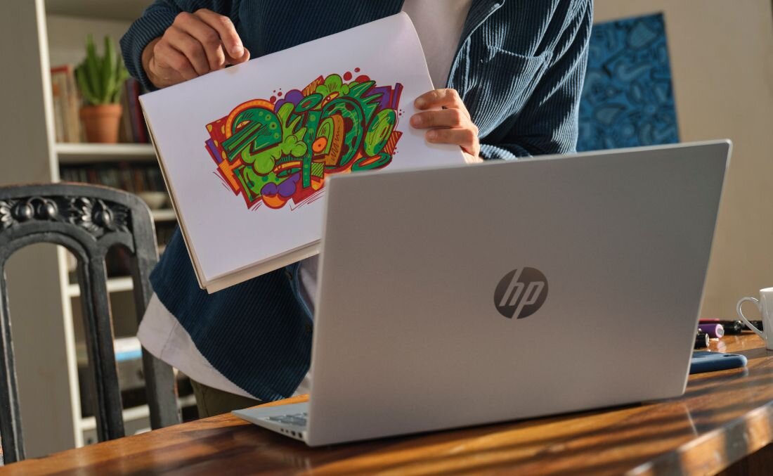 Kobieta w niebieskim swetrze pokazująca kolorową grafikę na kartce obok srebrnego laptopa HP OmniBook 5 16-BC1 na drewnianym stole - Pamięć LPDDR5 6400 MHz