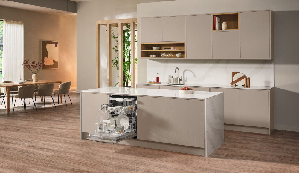 ZMYWARKA MIELE G 5662 SCVi Active S Zdjęcie przedstawia przestronną kuchnię z wyspą, w której zamontowana jest otwarta zmywarka wypełniona naczyniami. Jasna kolorystyka, eleganckie fronty meblowe i minimalistyczny układ nadają wnętrzu stylowy i funkcjonalny charakter