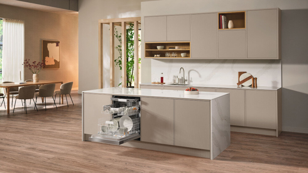 ZMYWARKA MIELE G 5811 SCi Active Plus CS Zdjęcie przedstawia przestronną kuchnię z wyspą, w której zamontowana jest otwarta zmywarka wypełniona naczyniami. Jasna kolorystyka, eleganckie fronty meblowe i minimalistyczny układ nadają wnętrzu stylowy i funkcjonalny charakter
