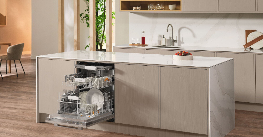 ZMYWARKA MIELE G 5862 SCVi Active Plus S Zdjęcie przedstawia przestronną kuchnię z wyspą, w której zamontowana jest otwarta zmywarka wypełniona naczyniami. Jasna kolorystyka, eleganckie fronty meblowe i minimalistyczny układ nadają wnętrzu stylowy i funkcjonalny charakter