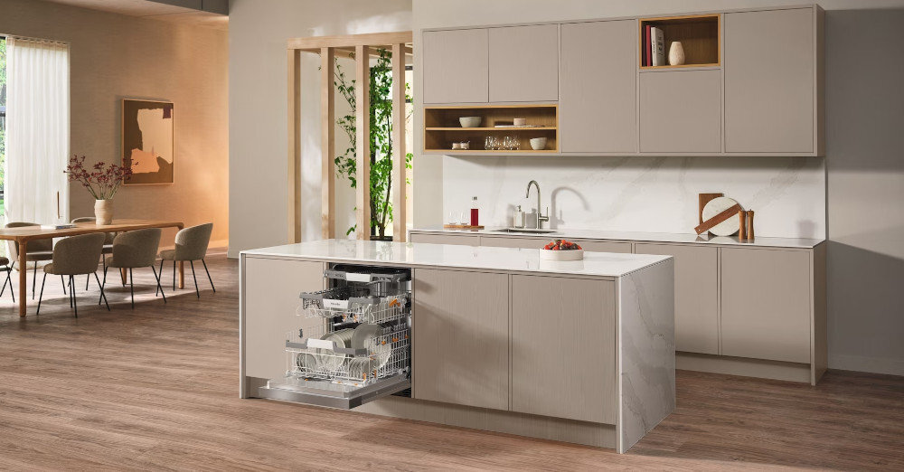 ZMYWARKA MIELE G 7040 SCi AutoDos CS Zdjęcie przedstawia przestronną kuchnię z wyspą, w której zamontowana jest otwarta zmywarka wypełniona naczyniami. Jasna kolorystyka, eleganckie fronty meblowe i minimalistyczny układ nadają wnętrzu stylowy i funkcjonalny charakter