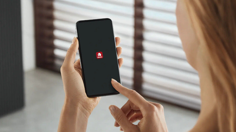 ZMYWARKA MIELE G 7040 SCi AutoDos CS Zdjęcie przedstawia osobę trzymającą smartfon, na którego ekranie widoczna jest ikona aplikacji Miele, symbolizująca możliwość zdalnego sterowania urządzeniami domowymi. Ujęcie podkreśla wygodę obsługi sprzętów za pomocą telefonu oraz charakter rozwiązania Miele@home inteligentny dom sterowanie aplikacją obsługa głosowa WLAN Miele Cloud integracja smart home