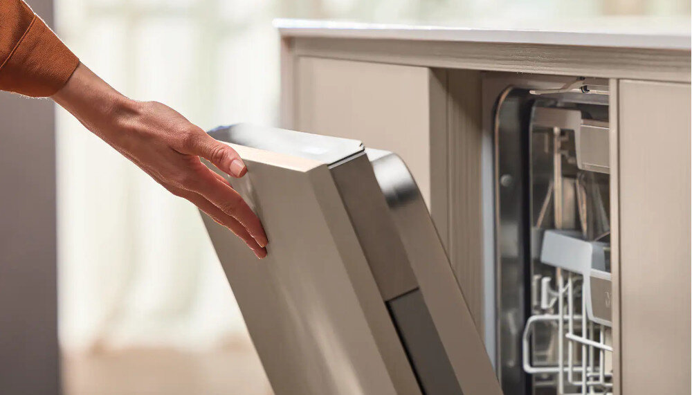 ZMYWARKA MIELE G7085 SCVI XXL Zdjęcie przedstawia osobę otwierającą zmywarkę w zabudowie, trzymając dłonią za krawędź frontu urządzenia. Ujęcie podkreśla wygodę użytkowania oraz płynny mechanizm otwierania drzwi zmywarki ComfortClose lekkie otwieranie regulowana pozycja ciche zamykanie