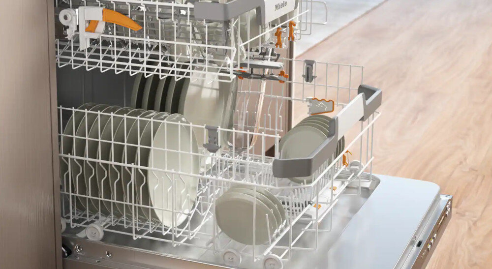 ZMYWARKA MIELE G7082 SCVI Zdjęcie przedstawia otwartą zmywarkę z wysuniętymi koszami, w których starannie ułożono talerze różnych rozmiarów. Widoczne pomarańczowe elementy regulacyjne podkreślają możliwość dostosowania przestrzeni, co ułatwia mycie naczyń o nietypowych kształtach 3D-MultiFlex szuflada na sztućce FlexCare Glass Bottle Multiclip XL-Assist elastyczny kosz stabilne kieliszki