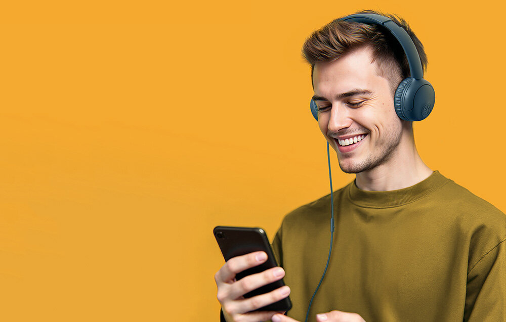 Słuchawki nauszne JLAB Studio 2 komfort Cloud Foam regulacja składana konstrukcja wygoda noszenia, Uśmiechnięty mężczyzna w turkusowych słuchawkach na uszach patrzący na ekran smartfona na żółtym tle