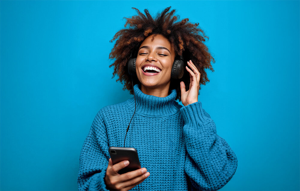 Słuchawki nauszne JLAB Studio 2 mikrofon pilot sterowanie rozmowy kontrola muzyki przewodowe, Kobieta z kręconymi włosami w niebieskim golfie i słuchawkach, śmiejąca się z telefonem w dłoni