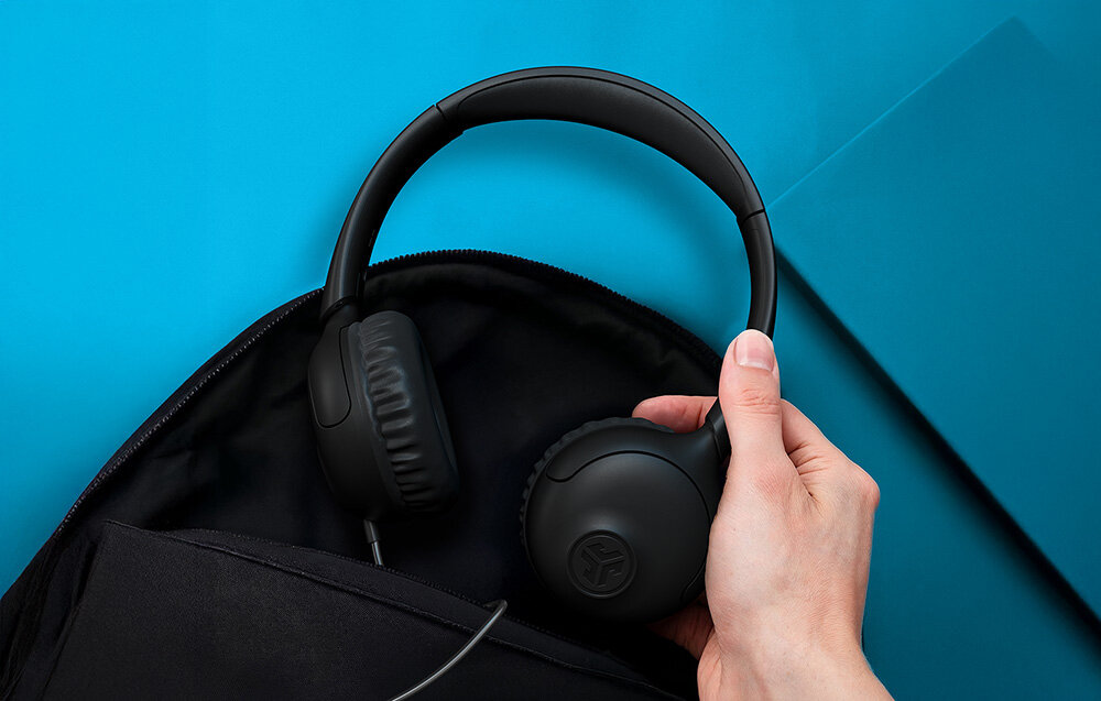 Słuchawki nauszne JLAB Studio 2 plug and play bez ładowania szybkie podłączenie przewodowe użytkowanie, Dłoń wkładająca czarne słuchawki nauszne do ciemnego plecaka na niebieskim tle