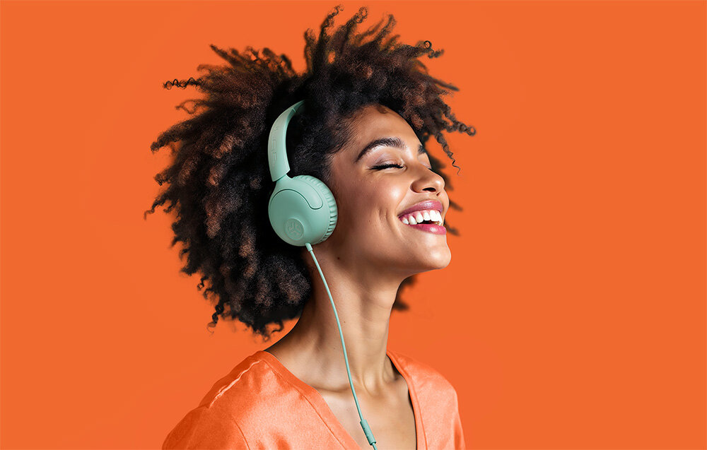 Słuchawki nauszne JLAB Studio 2 komfort Cloud Foam regulacja składana konstrukcja wygoda noszenia, Uśmiechnięta kobieta z kręconymi włosami w błękitnych słuchawkach na uszach na pomarańczowym tle