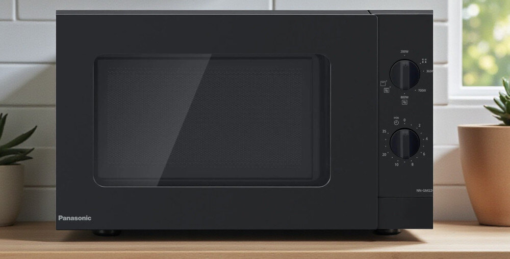 KUCHNIA MIKROFALOWA PANASONIC NN-GM22QBEPG Czarna mikrofalówka Panasonic ustawiona na blacie przy oknie, z dużą przeszkloną frontową szybą i prostym panelem z dwoma pokrętłami mocy i czasu po prawej stronie minimalistyczny design wygląd zaokrąglone krawędzie czytelne pokrętła matowe wykończenie drzwi estetyczne wykonanie codzienna wygoda stylowe AGD komfort użytkowania
