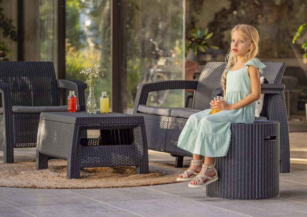 Stolik ogrodowy KETER Cool Stool 230905 z funkcją schowka Antracytowy dziecko siedzi na stoliku w tle inne meble ogrodowe dom i rośliny stabilność i komfort podczas siedzenia obciążenie do 110 kg solidne wykonanie dodatkowe miejsce do siedzenia
