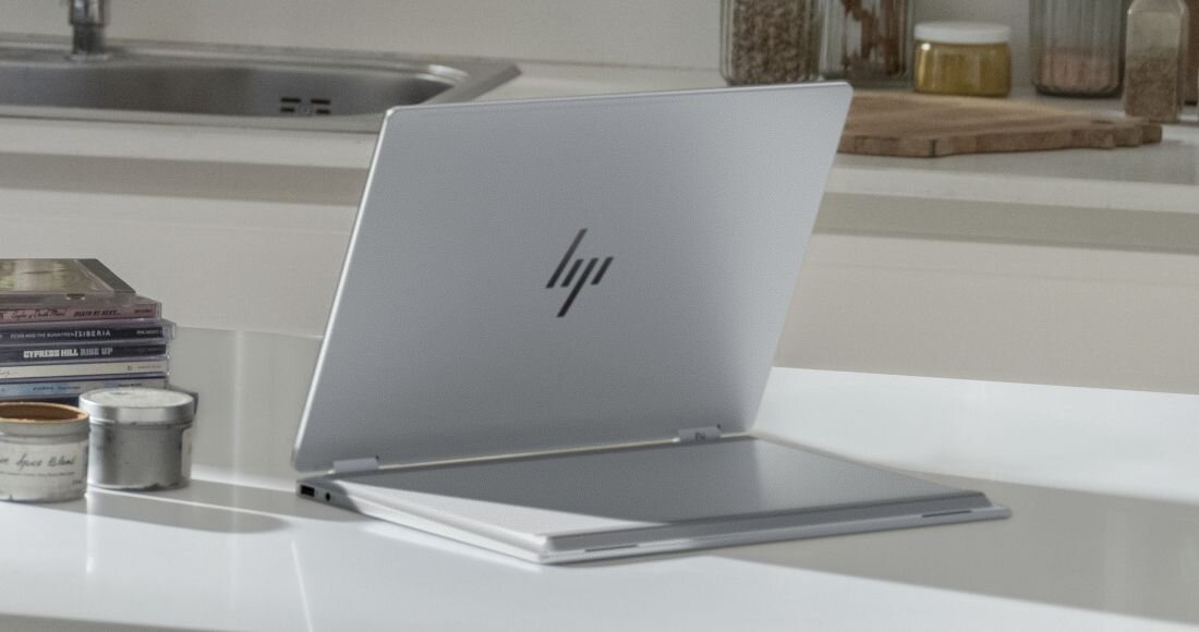 Srebrny laptop HP OmniBook rozłożony na blacie kuchennym, obok płyty CD i słoiczki z przyprawami. - metale pochodzące z recyklingu        