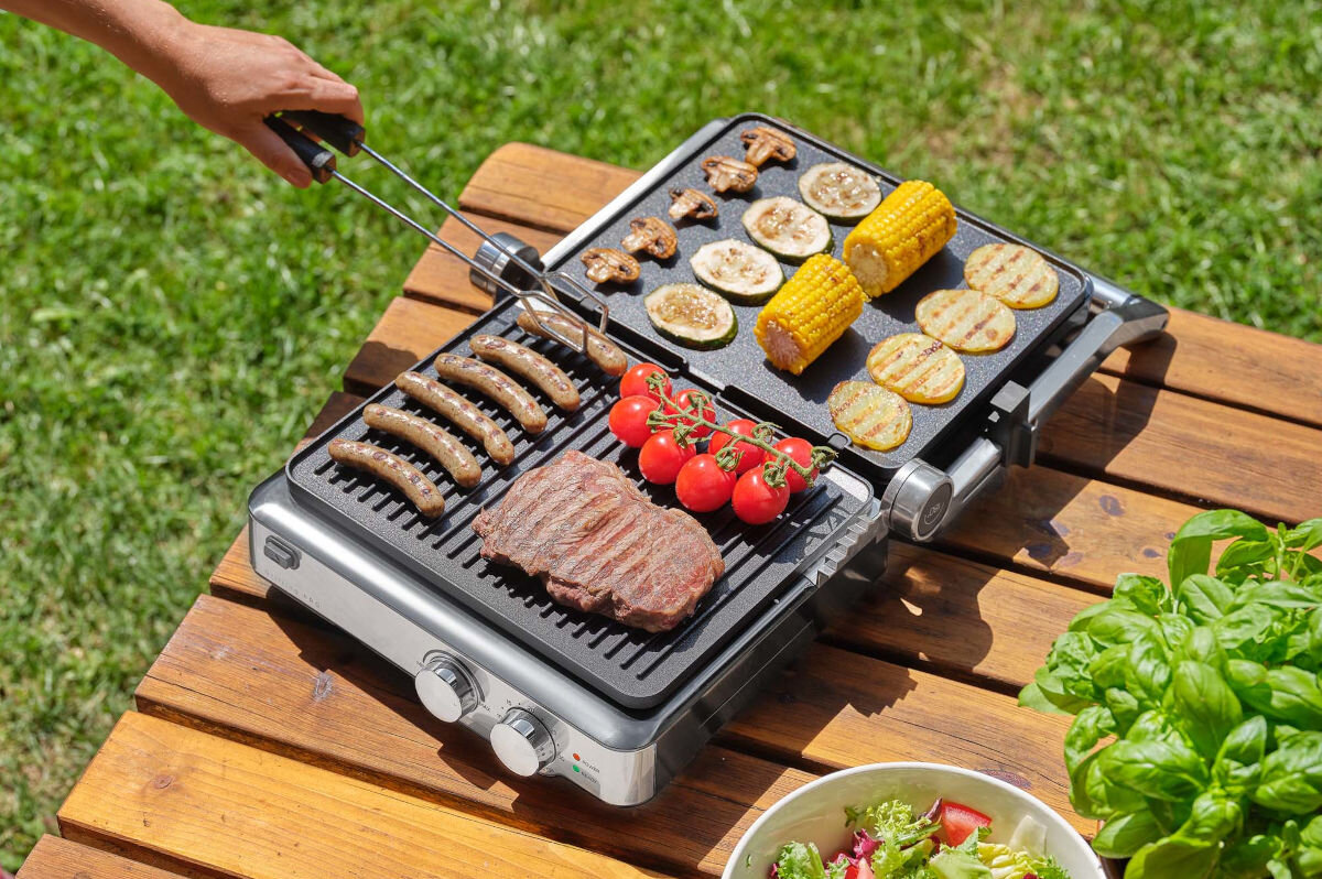 Grill elektryczny SENCOR SBG 4010SS, kuchnia, grillowanie, smażenie, potrawy, grill stoi na blacie drewnianego stołu w ogrodzie, w tle dłoń operuje szczypcami na potrawach grilla