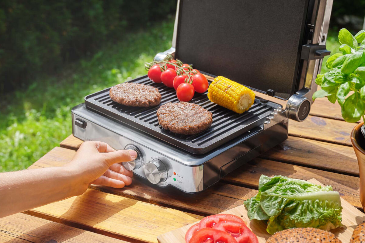 Grill elektryczny SENCOR SBG 4010SS, kolor, srebrno-czarny, style, industrialne, grill stoi na blacie drewnianym stołu w ogrodzie, dłoń operuje na pokrętłach grilla