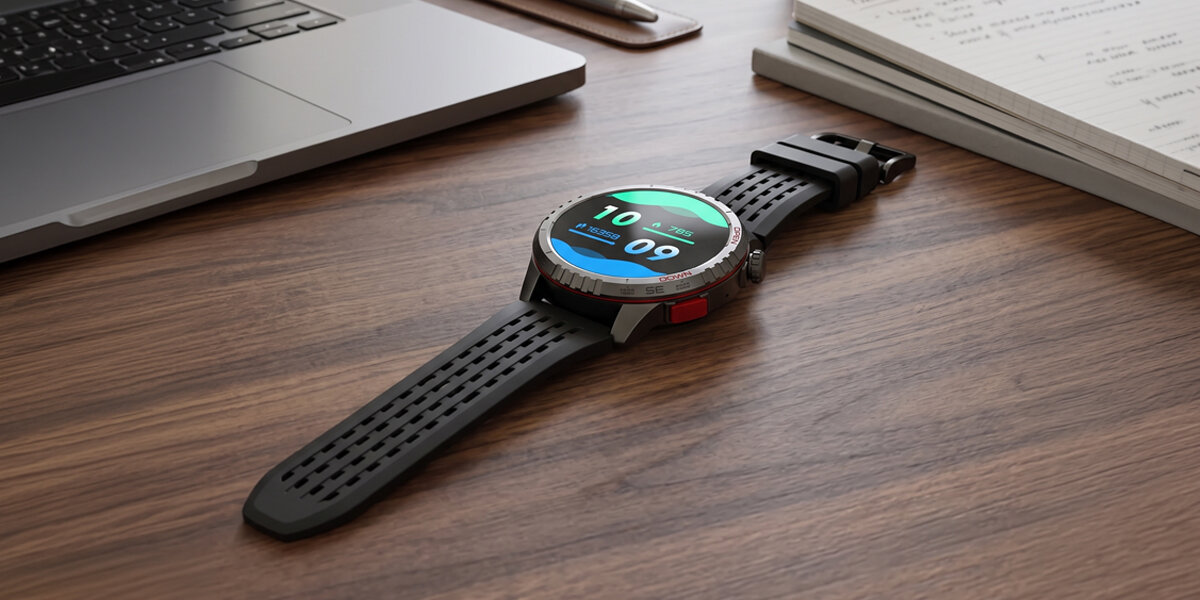Na drewnianym biurku leży czarny smartwatch z okrągłym wyświetlaczem, na którym widoczna jest godzina 10:09. Obok znajduje się otwarty laptop i notatnik.