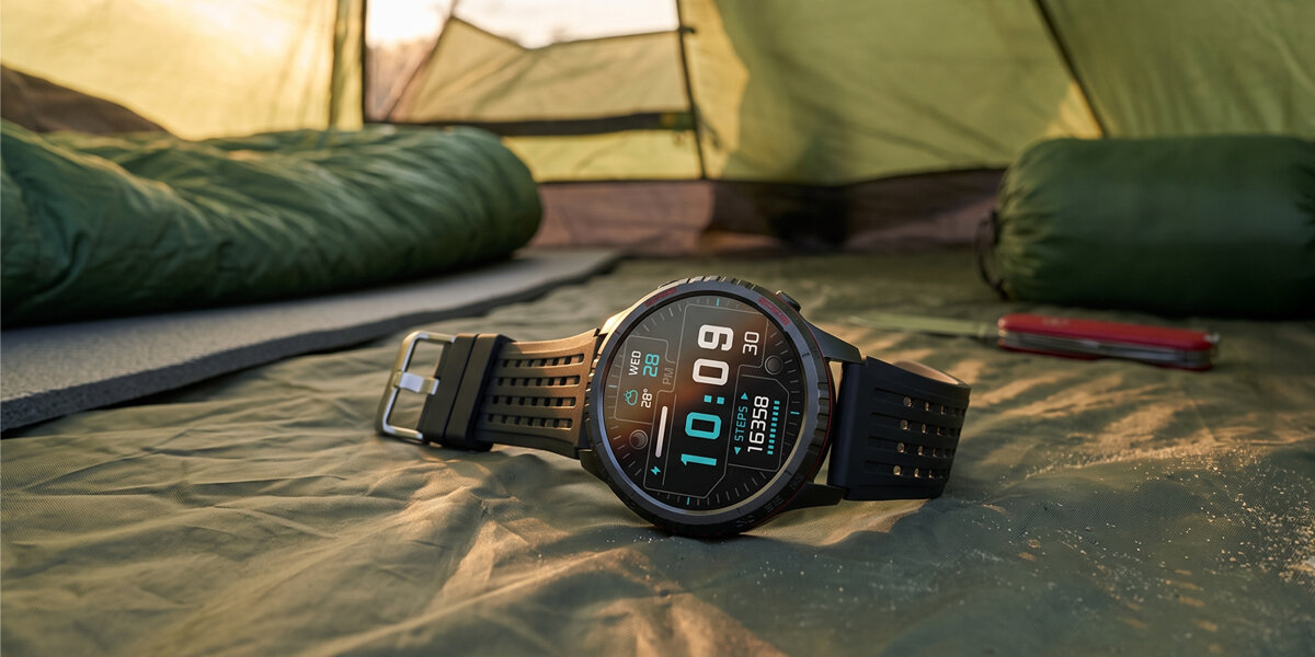 Smartwatch z czarnym paskiem leży na podłożu w namiocie. Na wyświetlaczu widoczna jest godzina 10:10 oraz liczba kroków 1538.