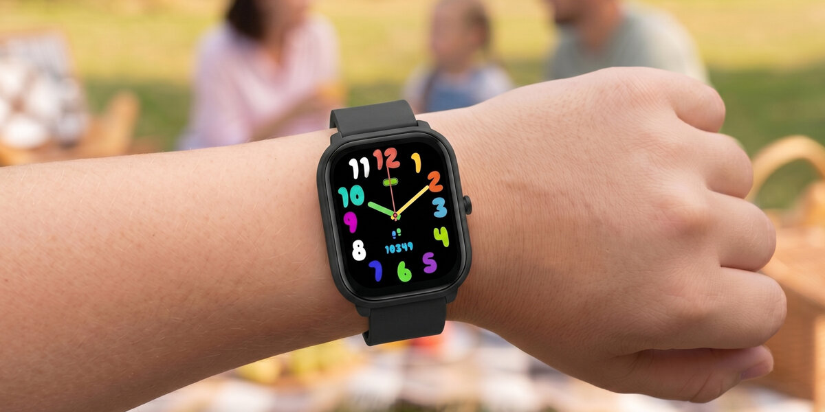 Zielony smartwatch Denver na nadgarstku dziecka podczas rodzinnego pikniku. Widoczna tarcza z zegarem z kolorowymi cyframi.