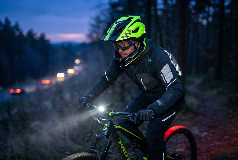 Osoba na rowerze górskim w lesie, nosząca zielony kask full face z odpinaną szczęką. W tle widać rozmyte światła samochodów na drodze.