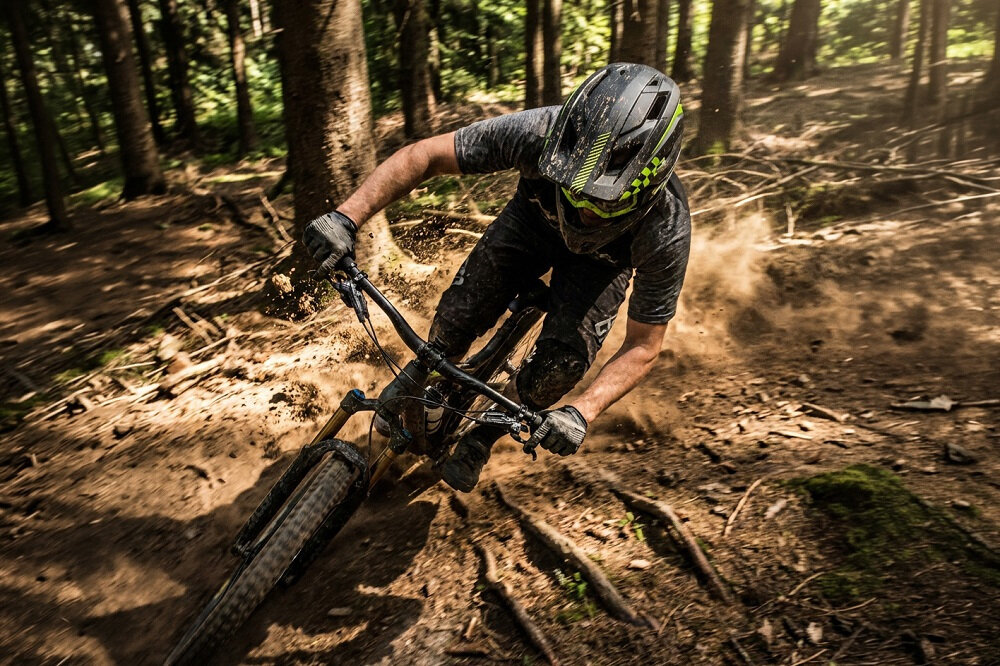 Osoba na rowerze górskim zjeżdża po leśnej ścieżce, nosząc czarny kask full face. W tle widać drzewa i unoszący się kurz.