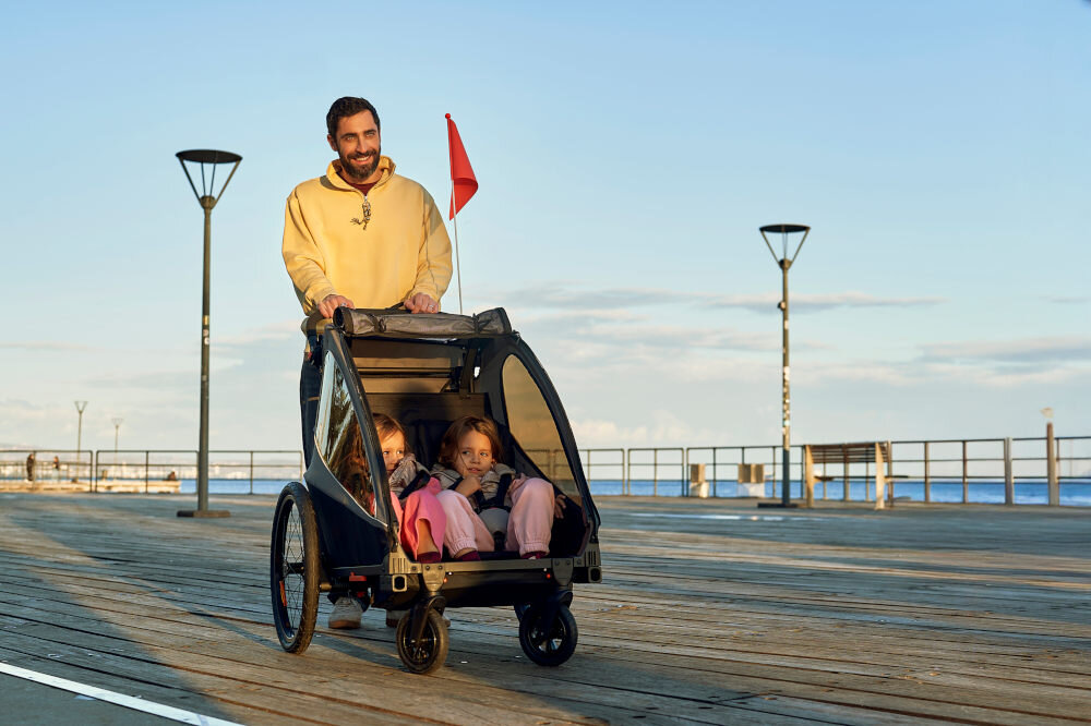 Przyczepka rowerowa KINDERKRAFT Nava Walk 2w1 60kg Pasy Czarno-szary przyczepka na białym tle widoczna z boku dyszel podłączony do koła rowerowego błyskawiczny montaż dzięki klikowym mocowaniom łatwe podłączenie do roweru możliwość przejazdu przez standardowe drzwi
